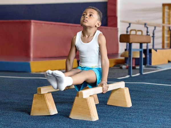 Picture of adorable dark skinned little gymnast competing on parallel bars. Hardworking talented African child exercising at gym, doing acrobatic moves, demonstrating strength, agility and flexibility